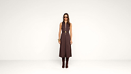 A woman stands against a plain white background, wearing a sleeveless brown dress with a belt, brown boots, large sunglasses, and gold bracelets.