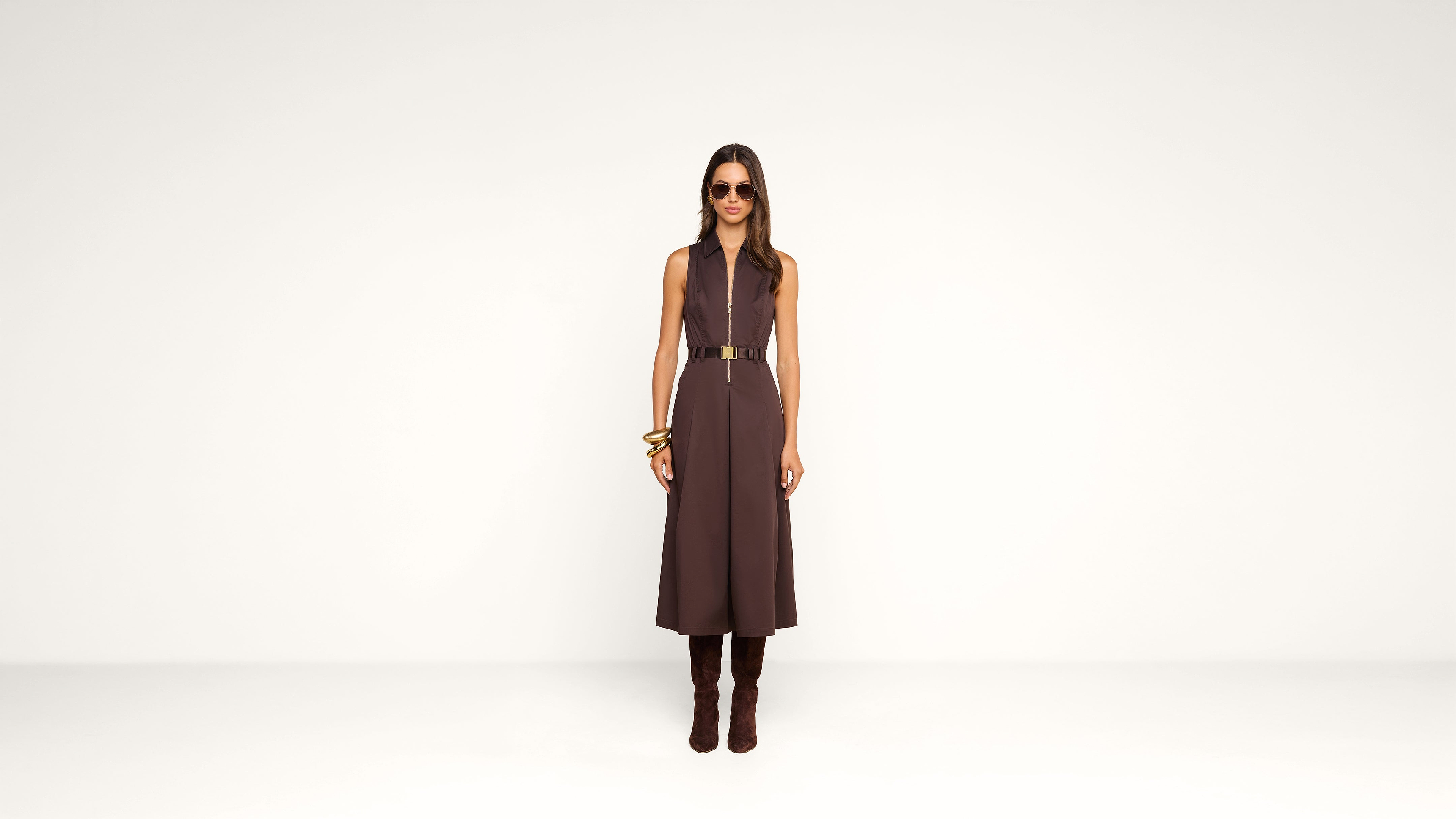 A woman stands against a plain white background, wearing a sleeveless brown dress with a belt, brown boots, large sunglasses, and gold bracelets.