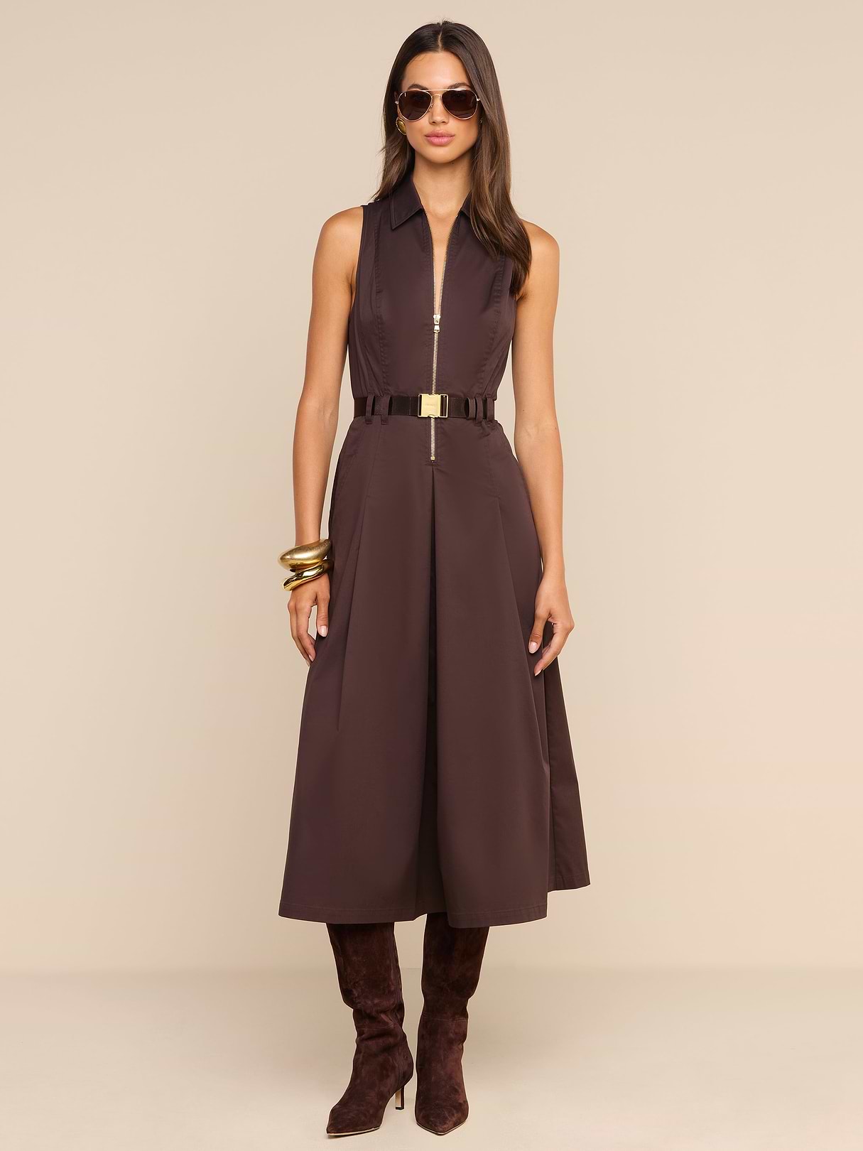 Woman wearing the Kolton Dress—a sleeveless brown cotton-blend poplin midi dress—styled with a gold belt, large gold bangles, dark brown boots, and sunglasses, standing against a plain white background.