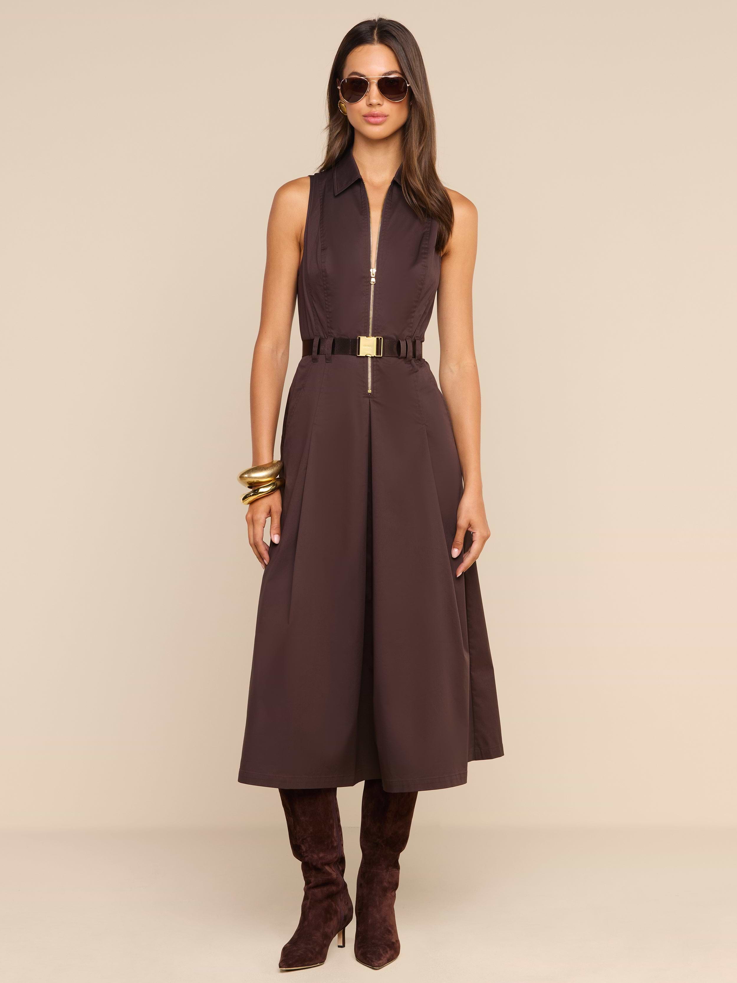 Woman wearing the Kolton Dress—a sleeveless brown cotton-blend poplin midi dress—styled with a gold belt, large gold bangles, dark brown boots, and sunglasses, standing against a plain white background.