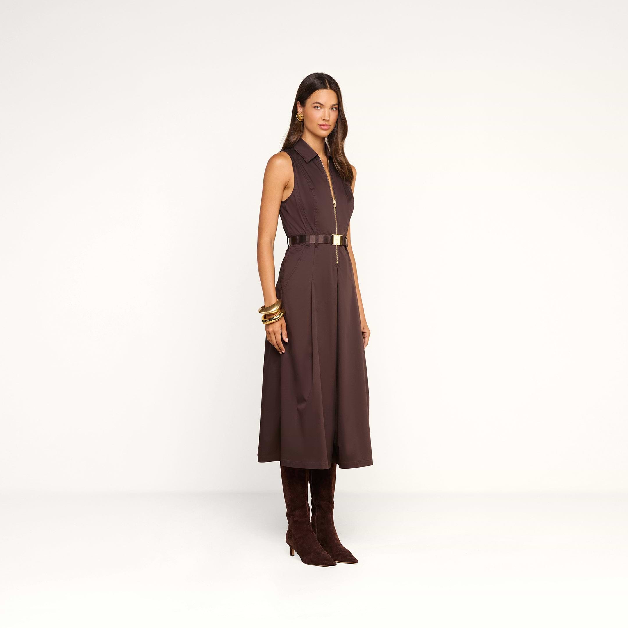 A woman stands against a plain white background wearing the Kolton Dress—a sleeveless, dark brown cotton-blend poplin midi dress—styled with gold bangles and knee-high brown boots.