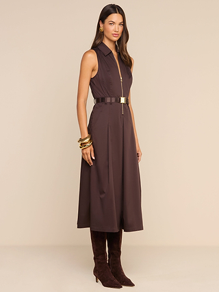 A woman stands against a plain beige background wearing the Kolton Dress—a sleeveless, belted brown center-zip midi dress—styled with gold hoop earrings, gold bangles, and knee-high brown suede boots for an effortless desk-to-drinks look.