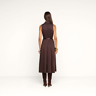 A woman stands facing away, showcasing the back of the Kolton Dress—a sleeveless brown cotton-blend poplin midi dress with a belt. She has long, straight hair and accessorizes with tall brown boots and gold bracelets. The background is plain white.