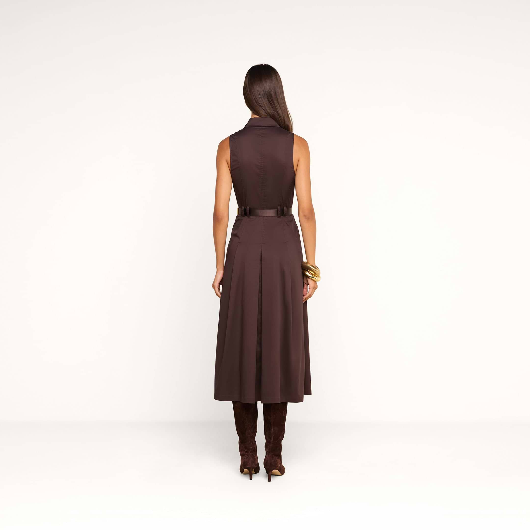 A woman stands facing away, showcasing the back of the Kolton Dress—a sleeveless brown cotton-blend poplin midi dress with a belt. She has long, straight hair and accessorizes with tall brown boots and gold bracelets. The background is plain white.