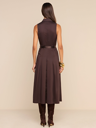 A woman stands facing away, wearing the sleeveless, dark brown Kolton Dress with a belt, tall brown boots, and gold bangles on her right wrist, against a plain beige background.