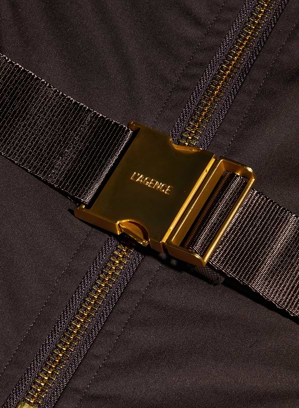 Close-up of a gold metal buckle labeled LAGENCE attached to a dark strap, placed diagonally over a black fabric with a visible gold zipper—an elegant detail featured on the Kolton Dress.