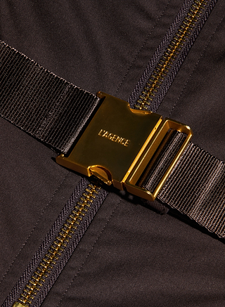 Close-up of a shiny gold buckle engraved with LAGENCE on a dark brown strap, placed over a brown fabric with a matching gold zipper — the perfect accent for your favorite Kolton Dress.