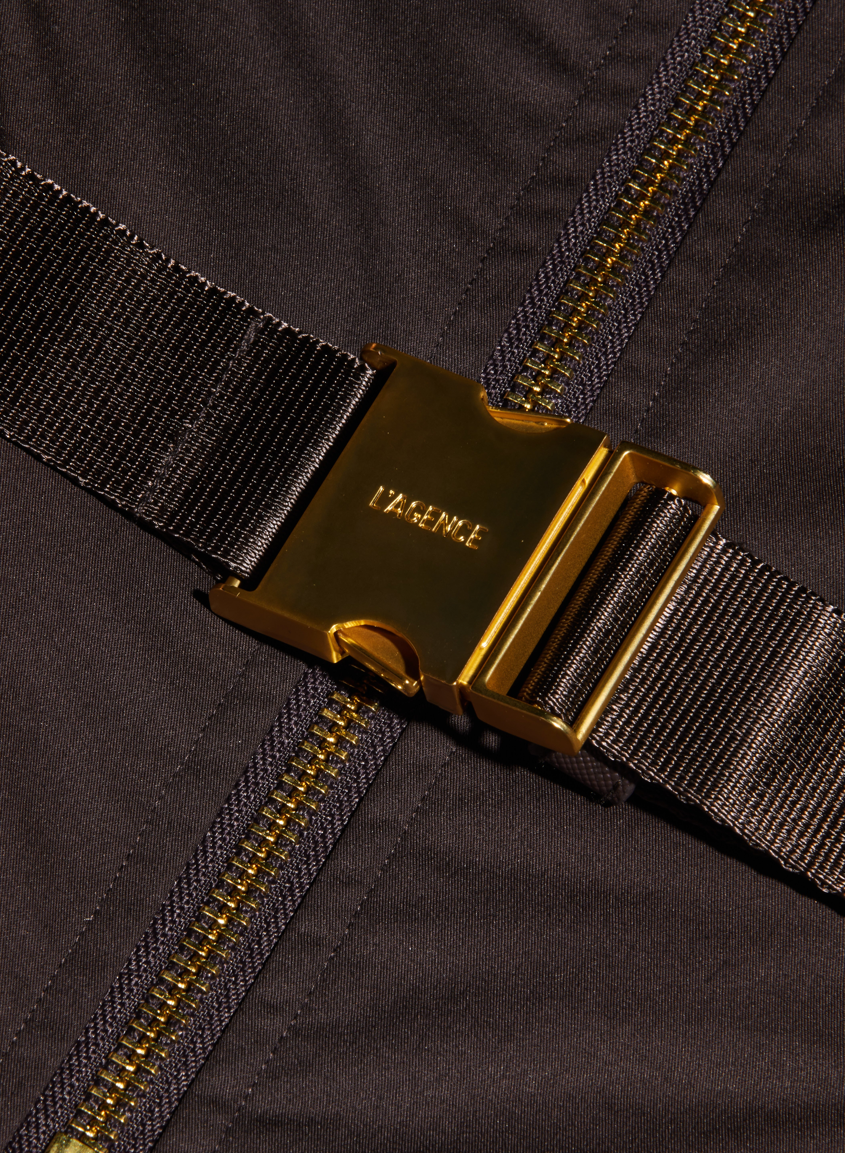 Close-up of a gold metal buckle labeled LAGENCE attached to a dark strap, placed diagonally over a black fabric with a visible gold zipper—an elegant detail featured on the Kolton Dress.