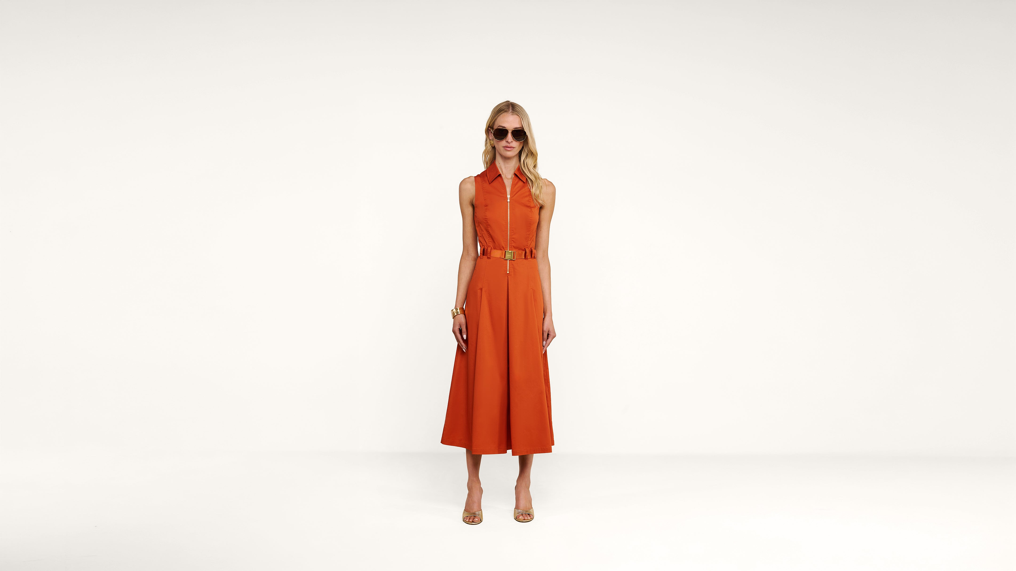 A woman stands against a white background wearing sunglasses and an orange sleeveless belted jumpsuit with wide legs, paired with beige heeled sandals.