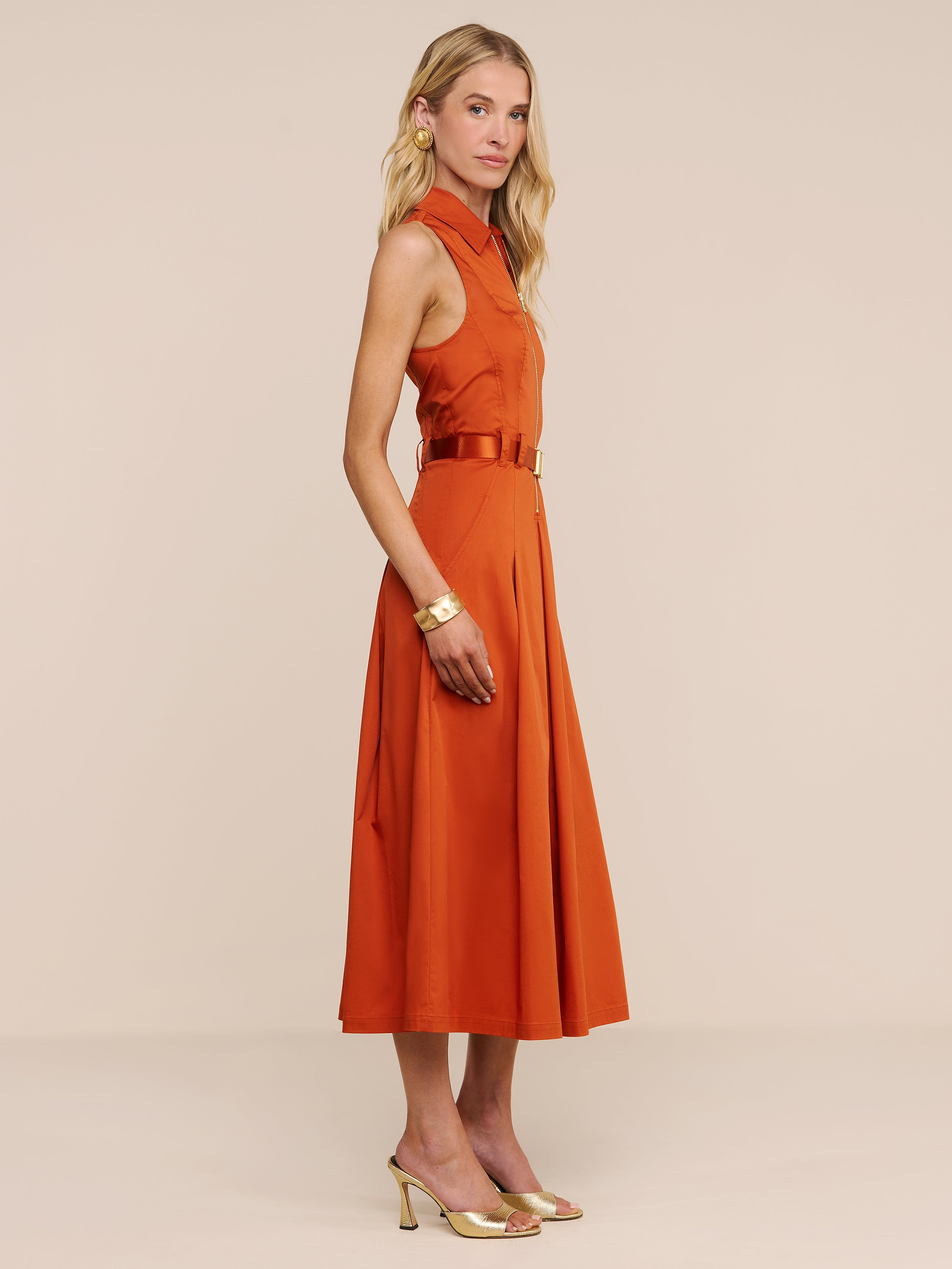 Una donna è ritratta di profilo, indossa un abito Kolton color arancio bruciato con cintura abbinata, orecchini a cerchio dorati, un braccialetto dorato e tacchi dorati — un look chic perfetto sia per l’ufficio che per un aperitivo — su uno sfondo beige a tinta unita.