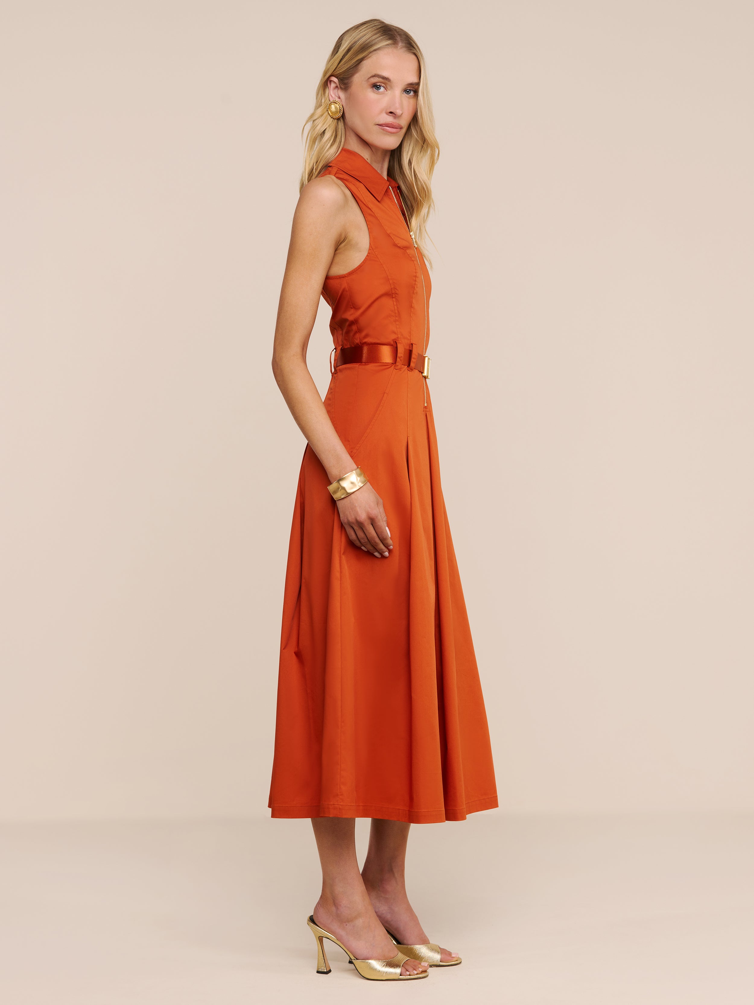 Una donna è ritratta di profilo, indossa un abito Kolton color arancio bruciato con cintura abbinata, orecchini a cerchio dorati, un braccialetto dorato e tacchi dorati — un look chic perfetto sia per l’ufficio che per un aperitivo — su uno sfondo beige a tinta unita.
