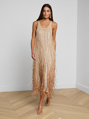 A woman poses indoors against a light wall and wooden floor, wearing the Prince Dress—a sleeveless, hand-embellished gold fringe midi dress—paired with strappy heels. She has long straight hair.