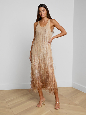 A woman stands in a minimal room wearing the Prince Dress, a sleeveless, tan, knee-length dress featuring hand-embellished crystal chain fringe. She poses with one hand on her hip and completes the look with strappy heeled sandals.