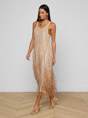 A woman wearing the sleeveless, beige Prince Dress with hand embellishments and delicate fringe, paired with strappy heeled sandals, walks indoors on a light wood floor against a plain white wall.
