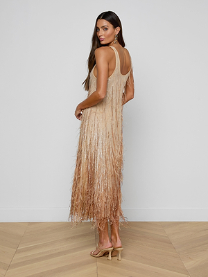 Woman with long brown hair wears the sleeveless Prince Dress—hand-embellished with fringe—and heeled sandals, standing on a light wood floor and looking over her shoulder against a white wall.