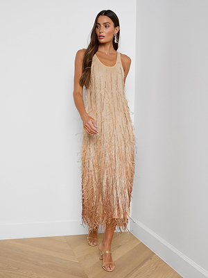 A woman with long, wavy brown hair stands in a minimal white room wearing the Prince Dress—gold, hand-embellished with crystal chain fringe—paired with matching strappy heels and hoop earrings.