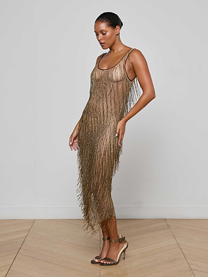 A woman poses on a wooden floor in the Gracelynn Beaded Fringe Dress, featuring a sleeveless design and antique gold bugle beads, paired with strappy high-heeled sandals against a plain light-colored wall.