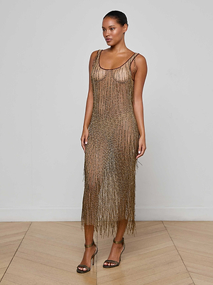A woman poses against a white wall in the Gracelynn Beaded Fringe Dress, adorned with antique gold bugle beads, a scoop neckline, and metallic high-heel sandals. One arm rests at her side while her other hand grazes her thigh.