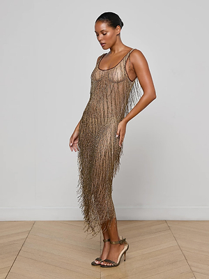 A woman in profile wears the Gracelynn Beaded Fringe Dress, detailed with antique gold bugle beads. She has slicked-back hair and high-heeled sandals, standing in a minimalist light-toned room with a wooden floor.