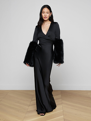 A woman in the Manon Gown with Faux Fur—a long black satin evening dress featuring a deep V-neckline and dramatic faux fur cuffs—walks on a light wooden floor against a plain white wall.
