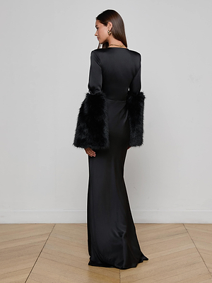 A woman stands on a wooden floor, facing away from the camera in front of a white wall, wearing the L'AGENCE Manon Gown with Faux Fur—an elegant, long fitted black satin dress featuring dramatic faux fur cuffs on the sleeves.