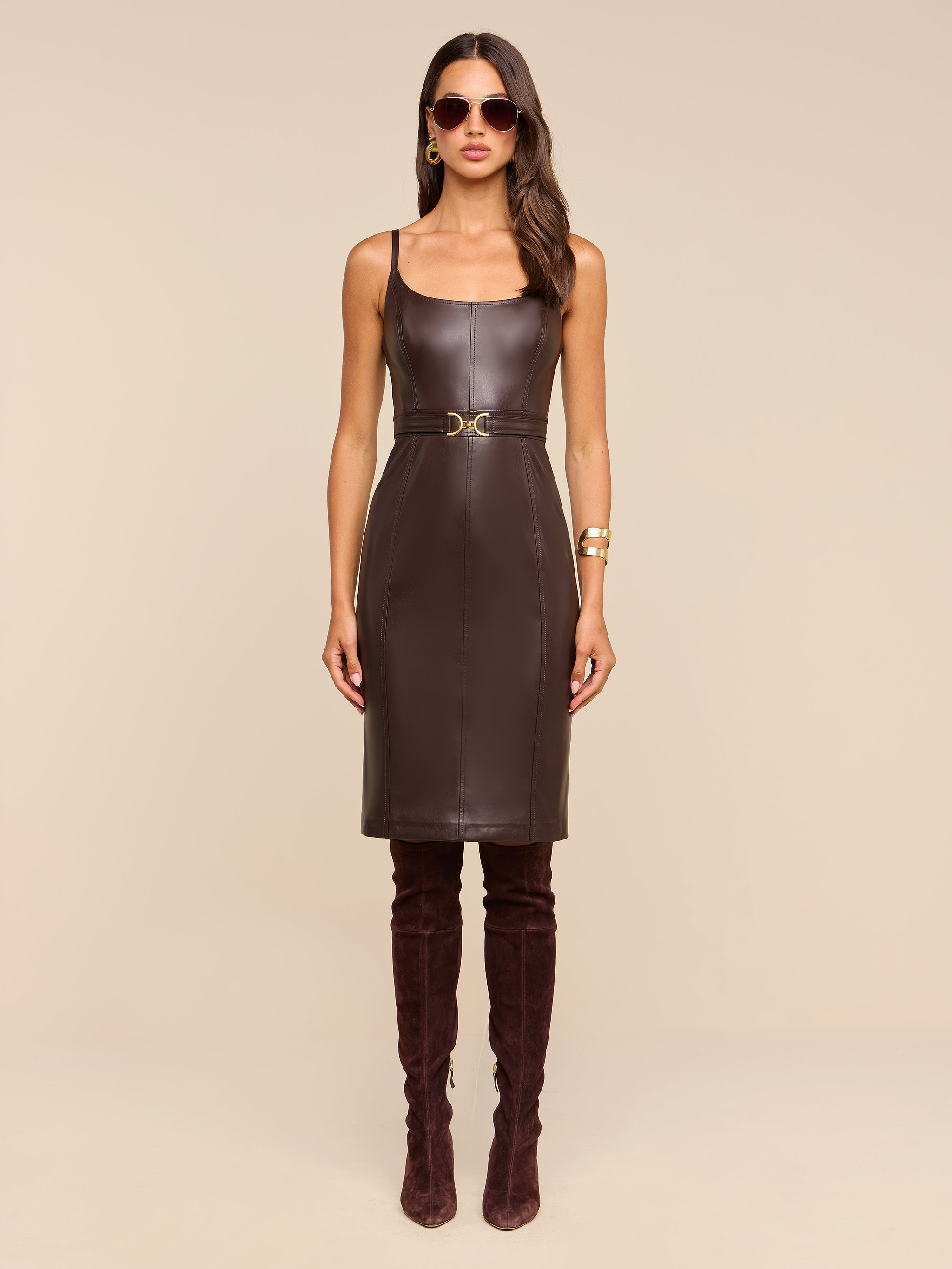 A woman wearing the Halia Vegan Leather Dress—sleeveless and brown with a gold buckle belt—paired with dark brown over-the-knee boots, sunglasses, hoop earrings, and a gold bracelet, stands against a plain beige background.