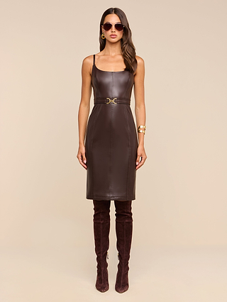 A woman wearing the Halia Vegan Leather Dress—sleeveless and brown with a gold buckle belt—paired with dark brown over-the-knee boots, sunglasses, hoop earrings, and a gold bracelet, stands against a plain beige background.