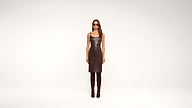 A woman stands against a plain white background wearing a sleeveless brown leather dress, tall brown boots, sunglasses, and gold bracelets.