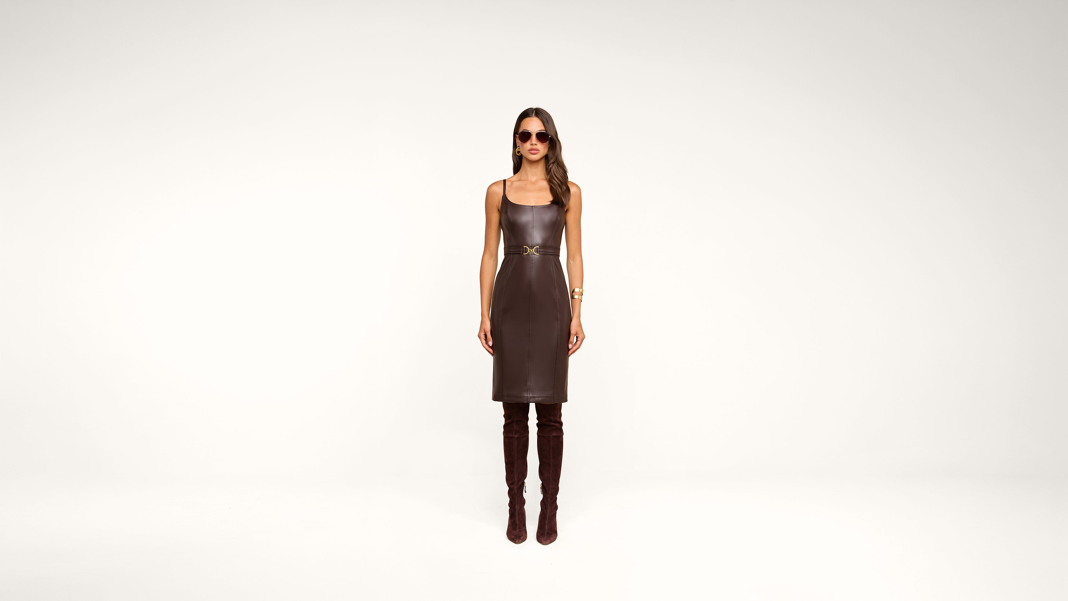 A woman stands against a plain white background wearing a sleeveless brown leather dress, tall brown boots, sunglasses, and gold bracelets.