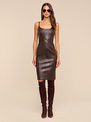 A woman wearing the Halia Vegan Leather Dress—a fitted brown midi dress with a gold buckle belt—styled with a gold bracelet, hoop earrings, sunglasses, and knee-high brown suede boots, standing against a beige background.
