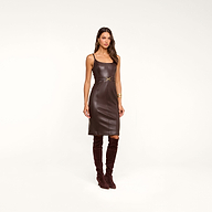 A woman wearing the Halia Vegan Leather Dress—a fitted, sleeveless style with a gold buckle belt at the waist—paired with brown knee-high boots, stands against a plain beige background.