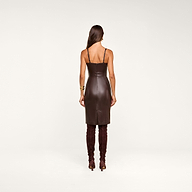 A woman stands facing away in the Halia Vegan Leather Dress, a fitted dark brown piece with thin straps and a gold buckle belt, styled with matching knee-high boots. She wears her long brown hair down and accessorizes with gold jewelry, set against a plain beige background.