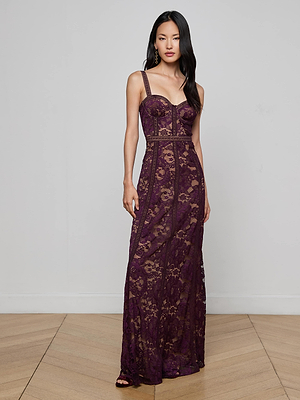 A woman with long, dark hair stands in a minimalist room wearing the Laynie Lace Maxi Dress—a deep purple, sleeveless floral Chantilly lace gown with a fitted bodice and sheer skirt—looking at the camera with a neutral expression.