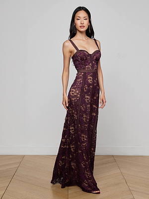 A woman with long dark hair poses against a light gray wall on a light wood floor, wearing the Laynie Lace Maxi Dress—a sleeveless, floor-length deep purple floral Chantilly lace dress with nude lining and thin straps.