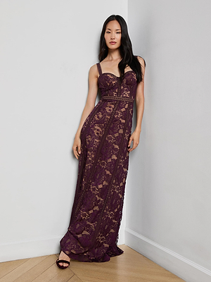 A woman with long dark hair stands against a white wall and wooden floor, wearing the Laynie Lace Maxi Dress—a floor-length, sleeveless deep purple floral Chantilly lace dress with sheer panels and strappy sandals.