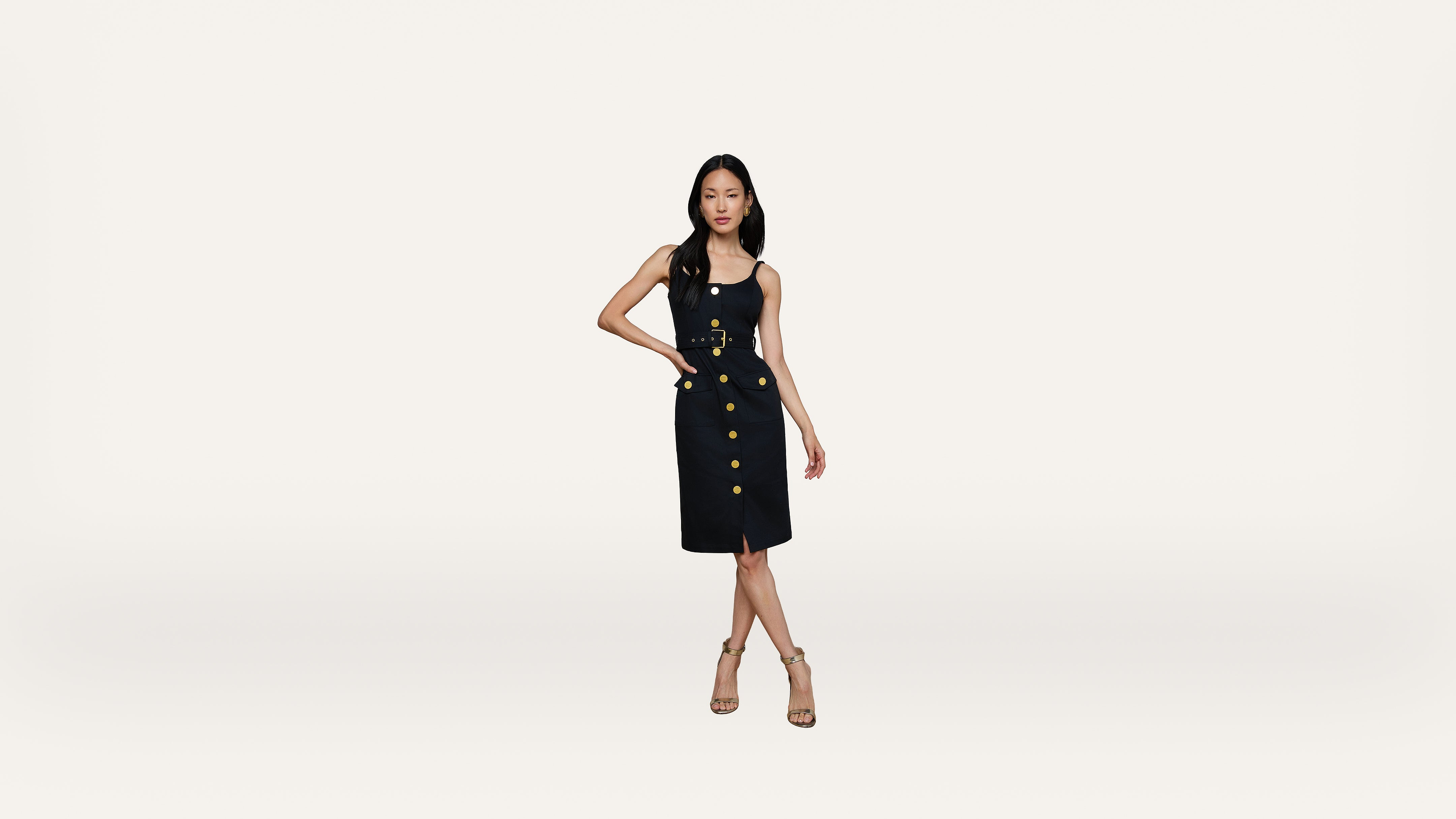 Woman wearing a sleeveless black dress with gold buttons and nude heels stands with one hand on her hip against a plain white background.