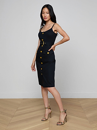 Wearing the Ida Dress—a sleeveless, gold button dress with a belt—a woman poses with one hand in her pocket and pairs the look with strappy heels against a plain white background.