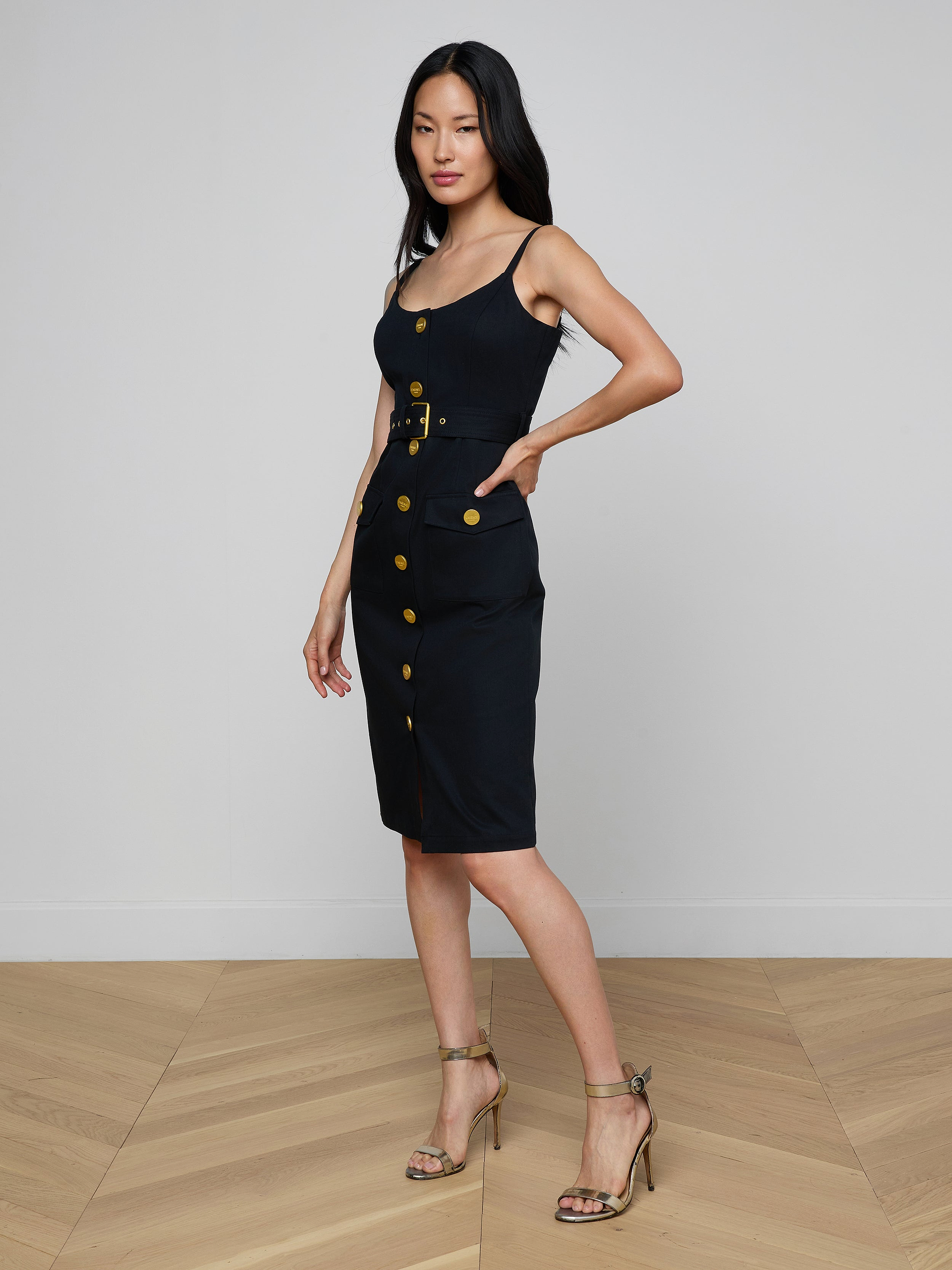 Wearing the Ida Dress—a sleeveless, gold button dress with a belt—a woman poses with one hand in her pocket and pairs the look with strappy heels against a plain white background.