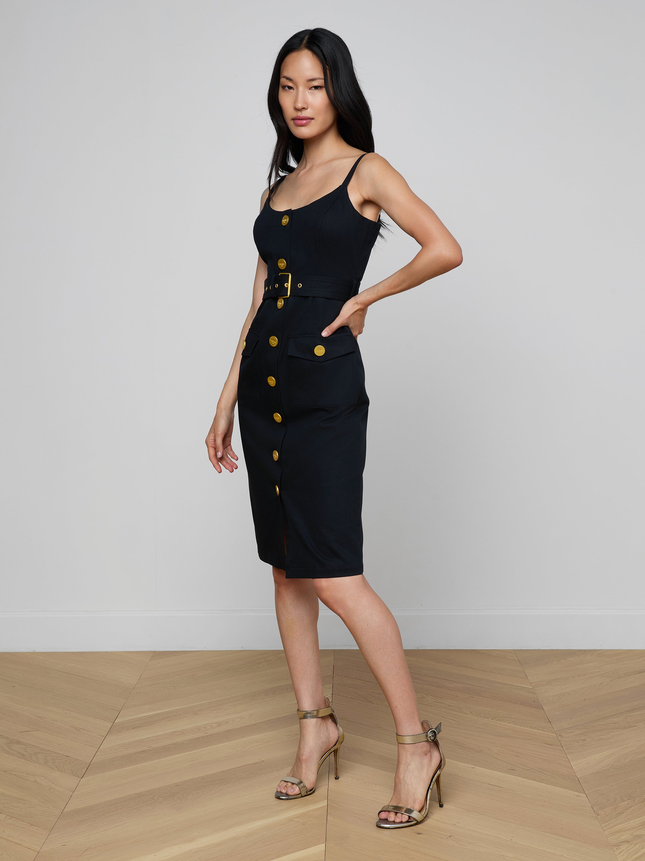 Wearing the Ida Dress—a sleeveless, gold button dress with a belt—a woman poses with one hand in her pocket and pairs the look with strappy heels against a plain white background.