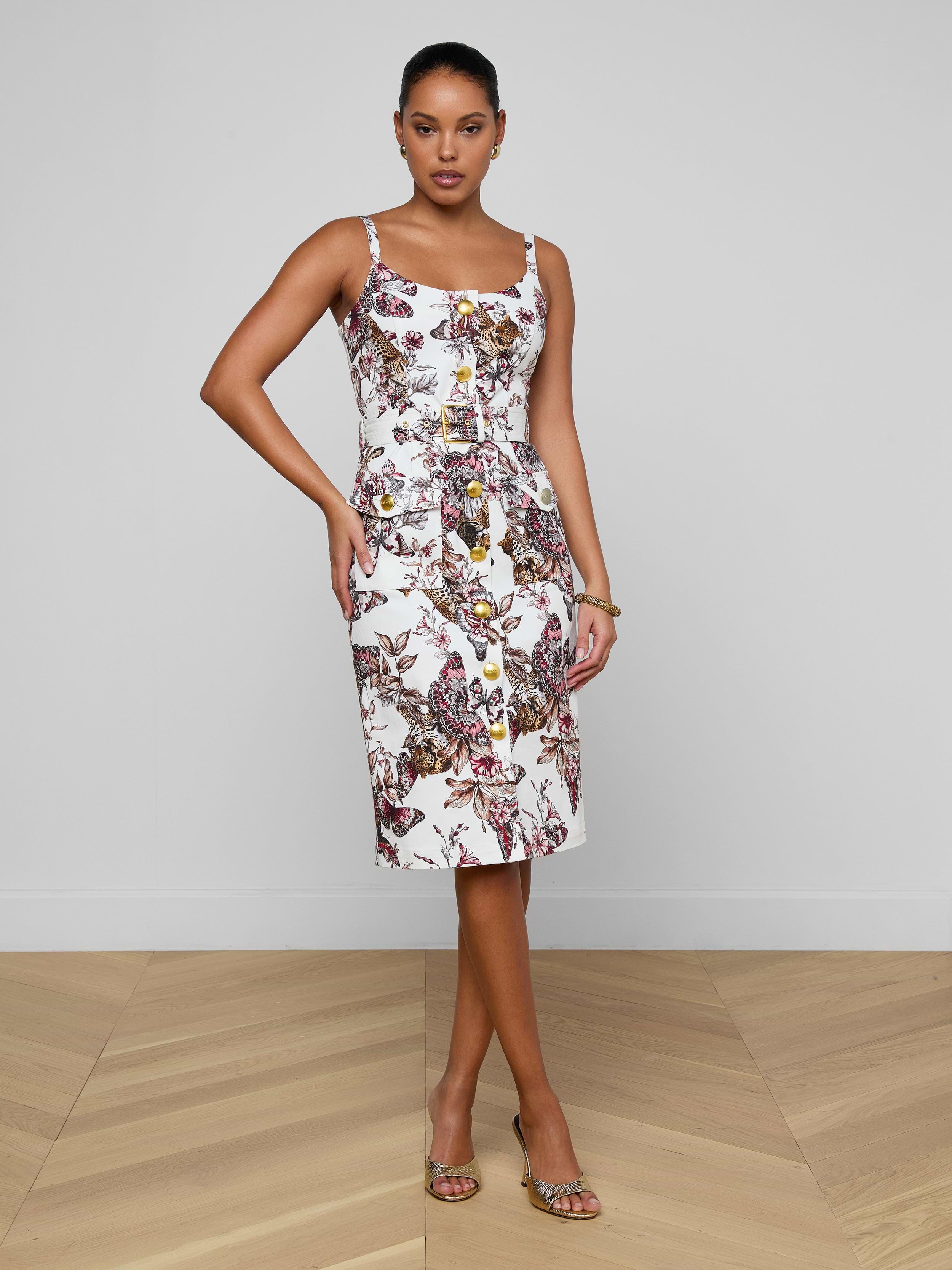 A woman poses against a plain white background wearing the Ida Dress—a sleeveless, knee-length dress featuring a vibrant butterfly jungle print, a fitted bodice, and a square neckline—styled with sandals and a bracelet.