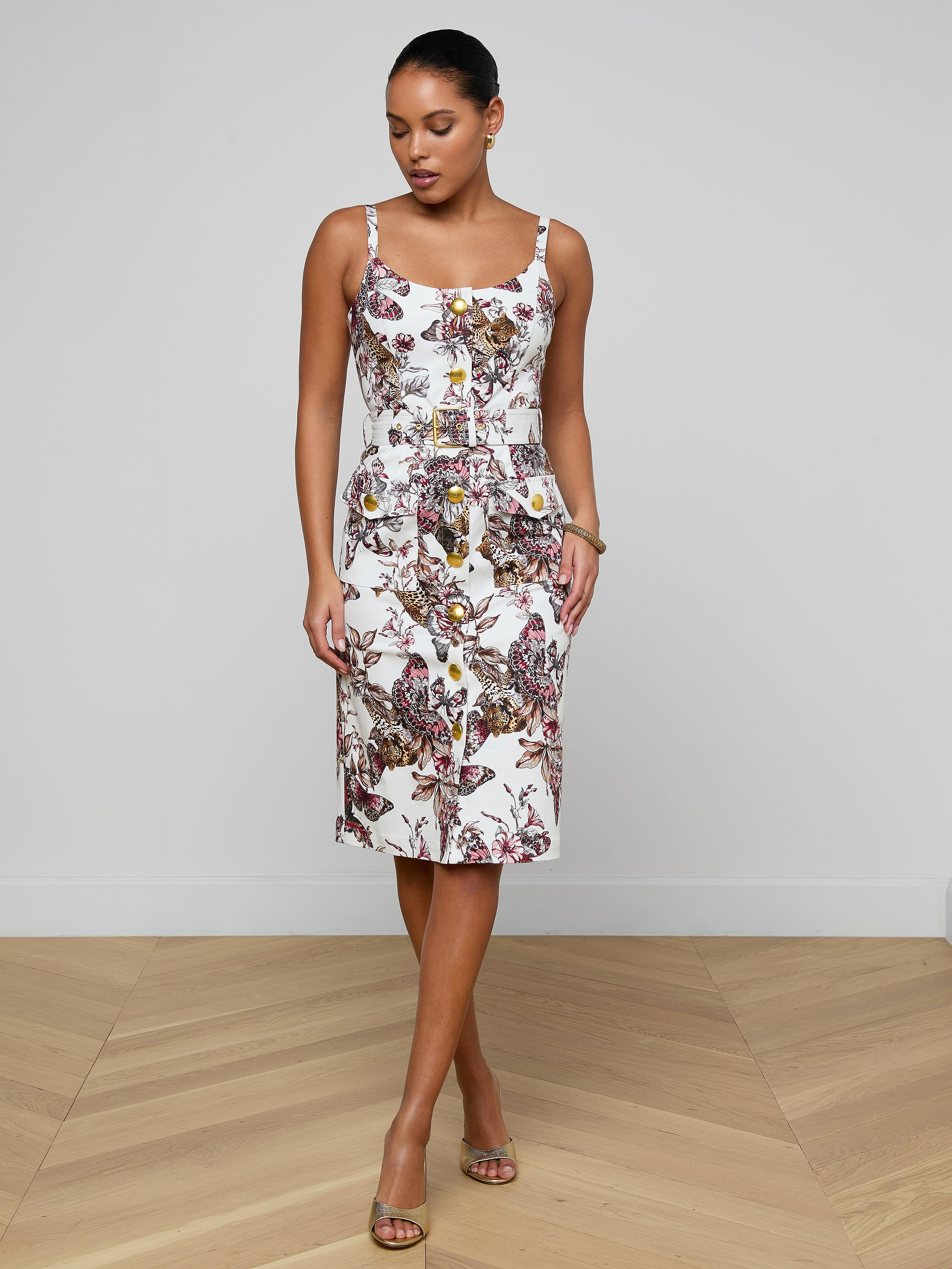 Wearing the Ida Dress—a sleeveless, fitted stretch cotton design adorned with a colorful floral pattern—a woman stands against a plain white background, looking down with her hands in her pockets.