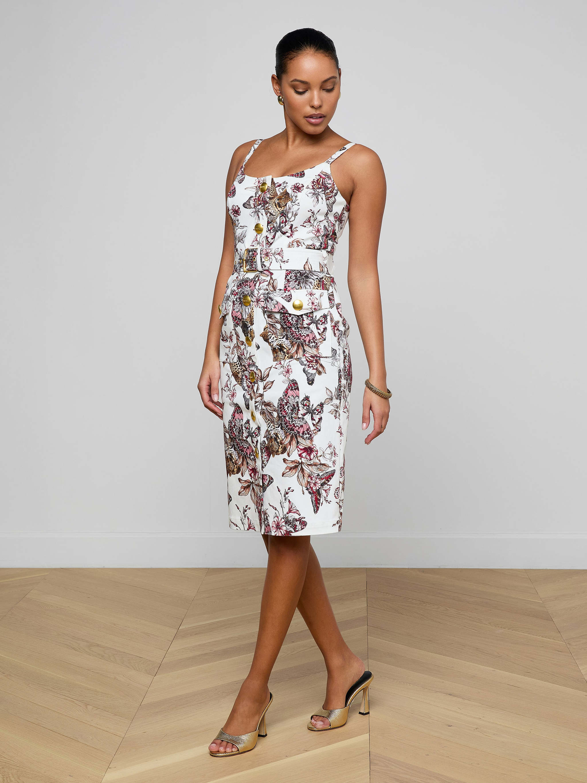 A woman wearing the Ida Dress—a white, sleeveless, knee-length stretch cotton dress with a colorful floral pattern—paired with tan high-heeled sandals, stands against a plain light background.