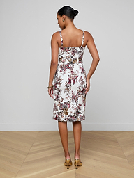 A woman stands facing away, wearing the Ida Dress—a tailored, sleeveless, knee-length stretch cotton dress adorned with a floral pattern—paired with beige heels. Her hair is styled in a neat bun against a plain, light-colored background.
