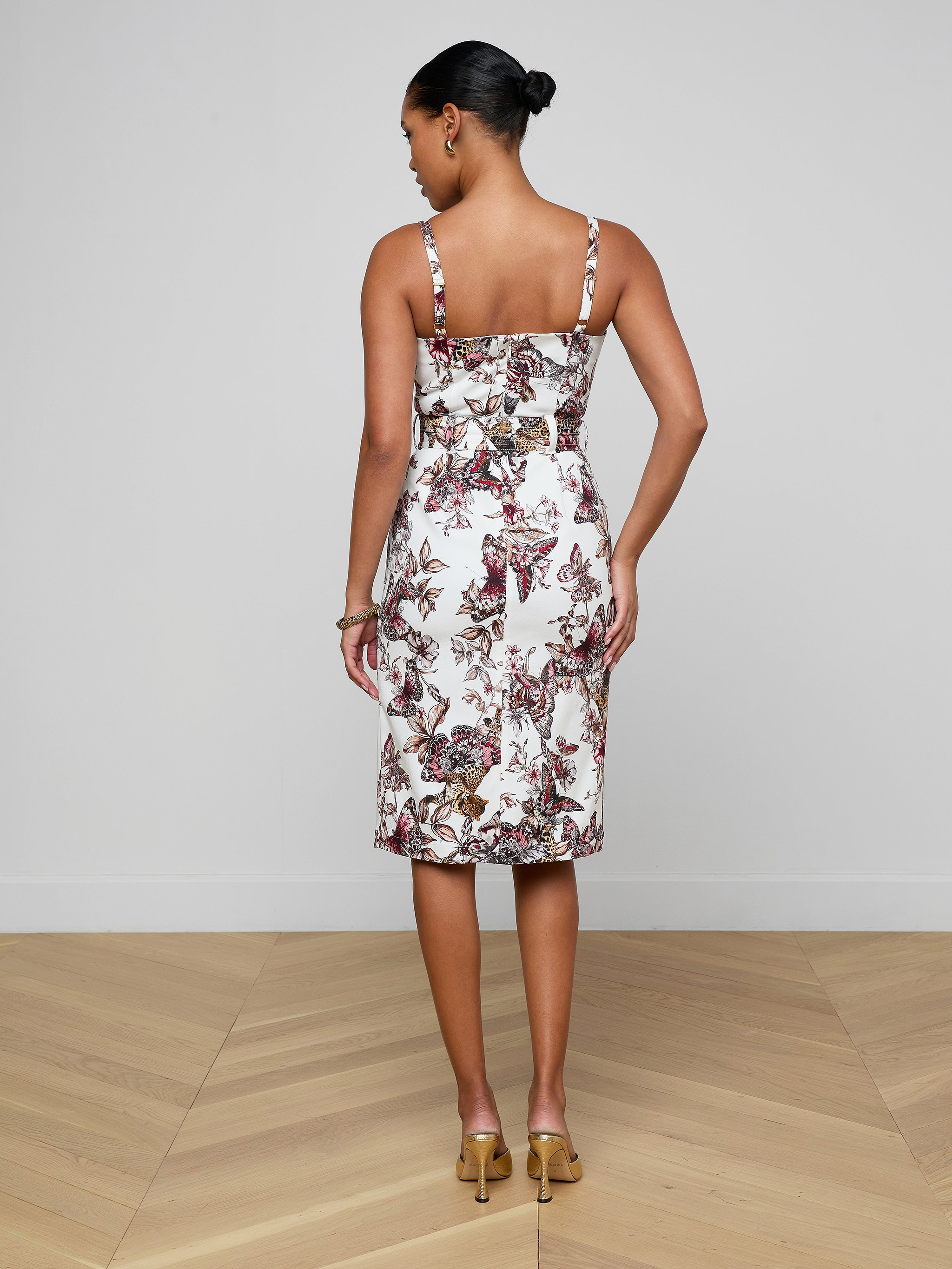 A woman stands facing away, wearing the Ida Dress—a tailored, sleeveless, knee-length stretch cotton dress adorned with a floral pattern—paired with beige heels. Her hair is styled in a neat bun against a plain, light-colored background.