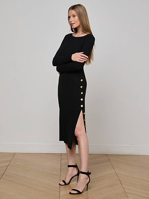 A woman with long, straight hair stands sideways in the L'AGENCE Astra Midi Dress, featuring a boat neckline, gold side-slit buttons, and a figure-flattering silhouette, paired with black heels on a wooden floor.