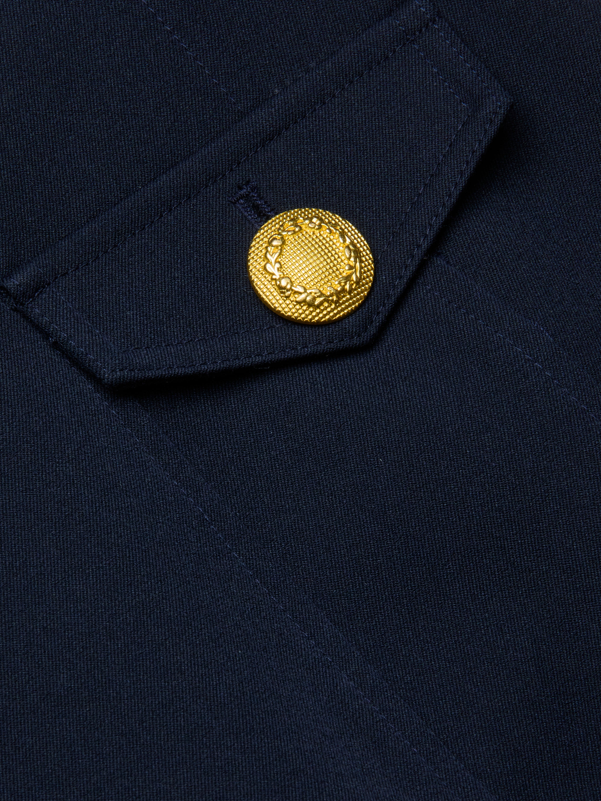 Close-up of the Cori Mini Dress in navy blue, showing a fabric pocket with a flap and gold-tone hardware—a decorative button embossed with a laurel wreath motif.