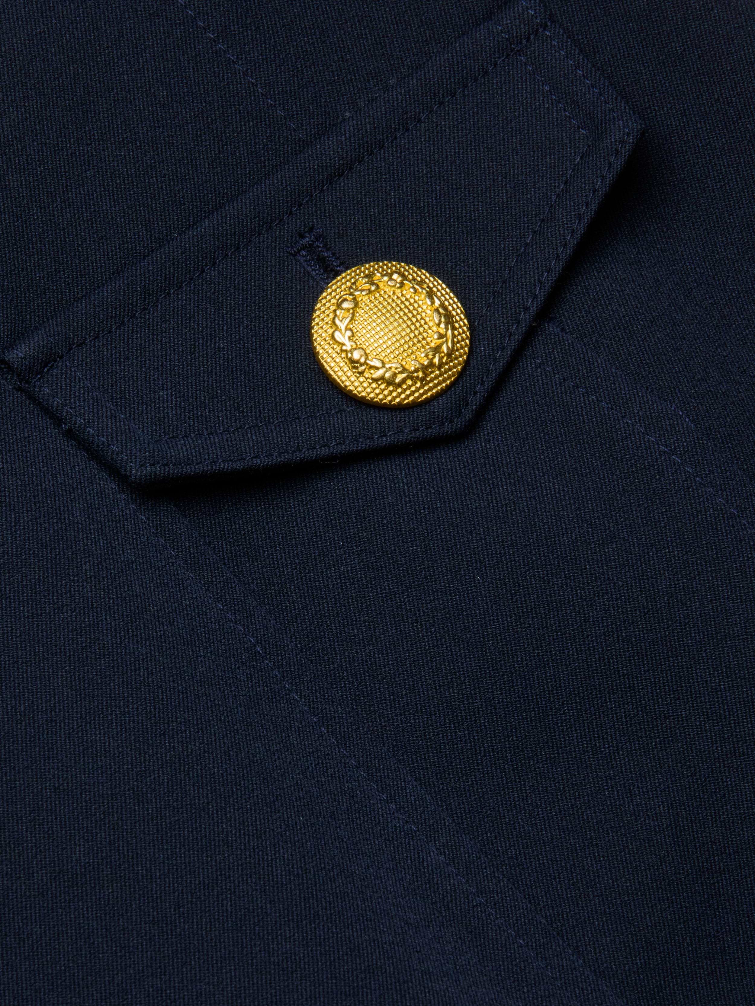 Close-up of the Cori Mini Dress in navy blue, showing a fabric pocket with a flap and gold-tone hardware—a decorative button embossed with a laurel wreath motif.