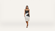 A woman stands against a plain white background, wearing a sleeveless white dress with black floral lace accents and black high-heeled sandals.