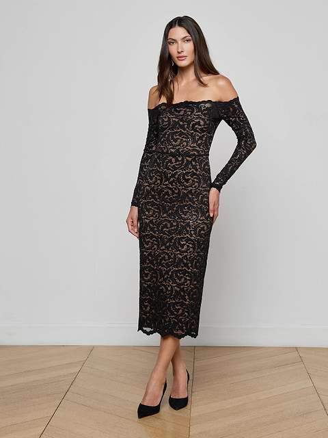 A woman wears the Lindsay Sequined Lace Dress, an off-the-shoulder black lace evening gown with long sleeves, paired with black heels, standing against a plain white background.