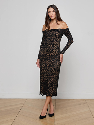 A woman poses on a wooden floor in the L’AGENCE Lindsay Sequined Lace Dress—an off-shoulder, black lace design with long sleeves and a fitted silhouette—paired with black heels against a plain white wall.